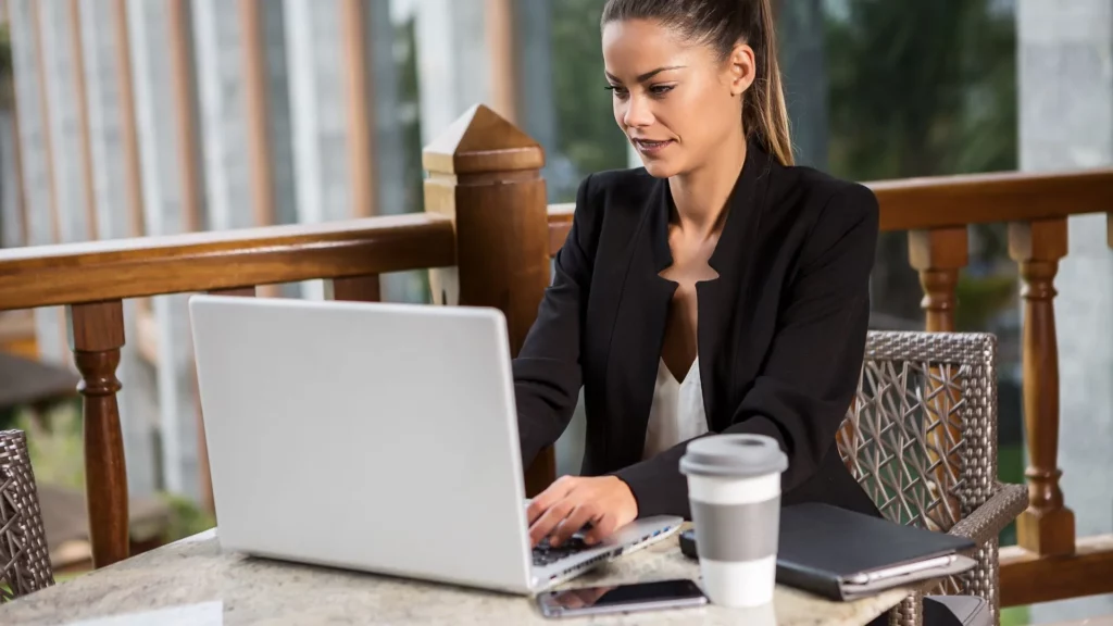 A woman dressed in business attire is typing on her laptop with her coffee and journal in front of her
