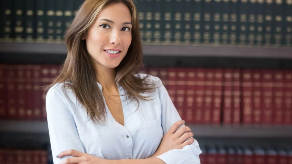 A female lawyer smiles to the camera with her arms crossed. Her background is a bookcase filled with law books.